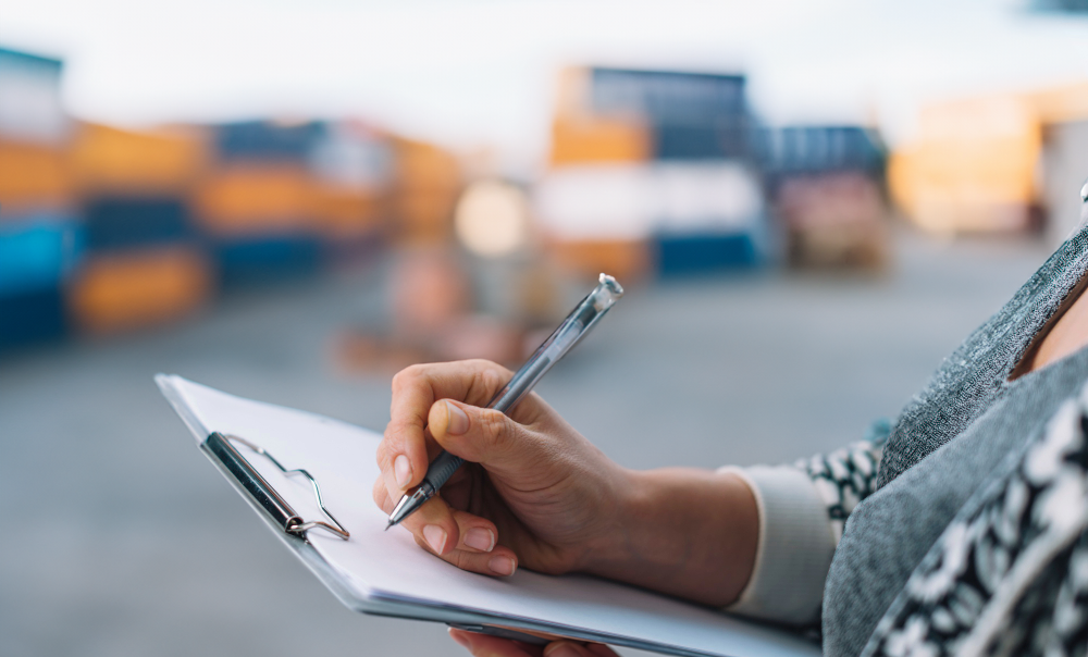 A woman in a shipping yard signing a piece of paper on a clip board